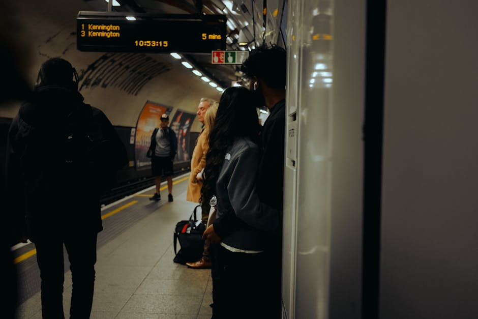 Inside an underground train station near Kennington, several passengers are waiting on the platform. The image shows a tiled floor with a smooth, polished surface, reflecting ambient lighting. A white pillar is positioned on the right side, and an electronic display board above indicates train times to Kennington station with bright yellow text on a black background. Passengers are dressed in casual clothing, some wearing face masks, standing near the platform edge. The scene is well-lit with ceiling lights, highlighting the clean, orderly environment. The overall setting suggests a typical station atmosphere, emphasizing hygiene and maintained surfaces, consistent with the professional cleaning standards of Kennington Carpet Cleaning, supporting the importance of surface cleanliness in public transit areas.