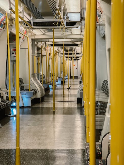 Inside an underground train station near Kennington, several passengers are waiting on the platform. The image shows a tiled floor with a smooth, polished surface, reflecting ambient lighting. A white pillar is positioned on the right side, and an electronic display board above indicates train times to Kennington station with bright yellow text on a black background. Passengers are dressed in casual clothing, some wearing face masks, standing near the platform edge. The scene is well-lit with ceiling lights, highlighting the clean, orderly environment. The overall setting suggests a typical station atmosphere, emphasizing hygiene and maintained surfaces, consistent with the professional cleaning standards of Kennington Carpet Cleaning, supporting the importance of surface cleanliness in public transit areas.