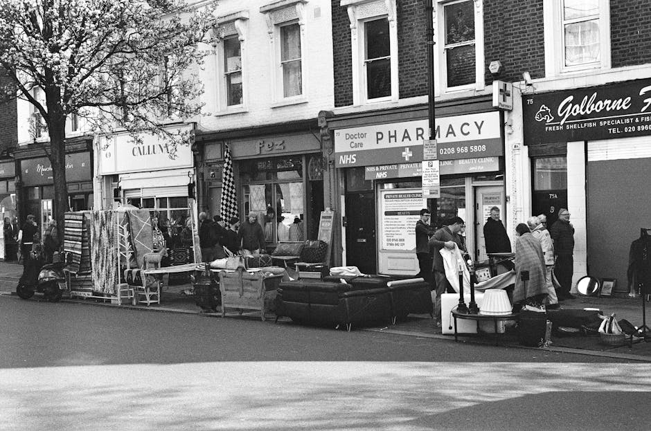 A black-and-white street scene on Kennington Road and SE11 streets showing a row of various shops including a pharmacy, fishmongers, and other retail outlets. The storefronts feature signage and large display windows with merchandise visible inside. In front of the shops, there are outdoor furniture pieces such as sofas, chairs, and tables arranged along the sidewalk, possibly for sale or part of a market setup. Several people are walking, browsing, or standing near the furniture, with some engaged in conversation or examining items. The scene includes a street sign on a pole in the middle distance, and mature trees are visible alongside the sidewalk, providing some shade. The overall appearance suggests a lively commercial area with a mix of pedestrians and street vendors, captured during daytime with natural lighting highlighting the various textures of the surfaces and displays.