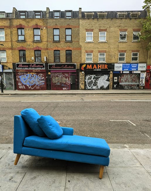 A bright blue chaise lounge with two matching cushions is placed on a concrete sidewalk, facing a busy street with graffiti-covered shopfronts and a row of residential brick buildings above. The lounge appears clean and well-maintained, contrasting with the urban backdrop. In the background, the shopfronts are closed, with graffiti tags and posters partially obscuring the shutters. The scene is illuminated by natural daylight, highlighting the vibrant color of the furniture against the muted tones of the buildings and street. This image exemplifies outdoor domestic furniture situated in an urban environment, with no visible cleaning equipment or products. For professional surface cleaning and deep cleaning of similar furniture, Kennington Carpet Cleaning offers expert services, as seen on their website kenningtoncarpetcleaning.org.uk.
