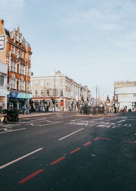 A black-and-white street scene on Kennington Road and SE11 streets showing a row of various shops including a pharmacy, fishmongers, and other retail outlets. The storefronts feature signage and large display windows with merchandise visible inside. In front of the shops, there are outdoor furniture pieces such as sofas, chairs, and tables arranged along the sidewalk, possibly for sale or part of a market setup. Several people are walking, browsing, or standing near the furniture, with some engaged in conversation or examining items. The scene includes a street sign on a pole in the middle distance, and mature trees are visible alongside the sidewalk, providing some shade. The overall appearance suggests a lively commercial area with a mix of pedestrians and street vendors, captured during daytime with natural lighting highlighting the various textures of the surfaces and displays.