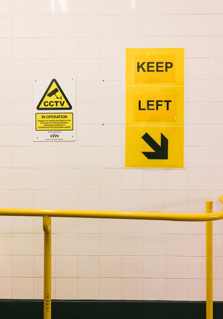 Interior view of a sterile, tiled corridor featuring a white wall with a CCTV warning sign and three yellow directional signs that read 'KEEP', 'LEFT', and an arrow pointing downward, indicating a controlled pathway. A yellow safety barrier runs horizontally across the lower part of the image. The clean, glossy tiles and bright lighting suggest a hygienic environment, suitable for thorough surface cleaning and sanitisation undertaken by Kennington Carpet Cleaning to address common access issues in flat communal areas.