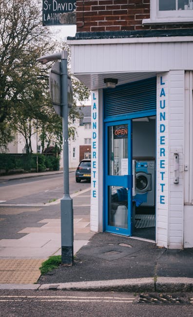 Exterior view of a small laundry shop situated on a street corner in Kennington. The shop has white wooden paneling and blue signage with the word 'LAUNDETTE' painted vertically on both sides of the entrance. The entrance features a glass door with a blue frame, through which a commercial washing machine is visible inside. A small 'OPEN' sign is lit in red above the door. Adjacent to the shop is a grey lamppost, and the sidewalk appears clean and well-maintained, with a tactile paving strip nearby. In the background, a street with parked cars and trees under natural daylight can be seen. This image highlights the external appearance of a typical laundrette, emphasizing cleanliness and accessibility in a residential area, with no visible cleaning tools or surfaces.