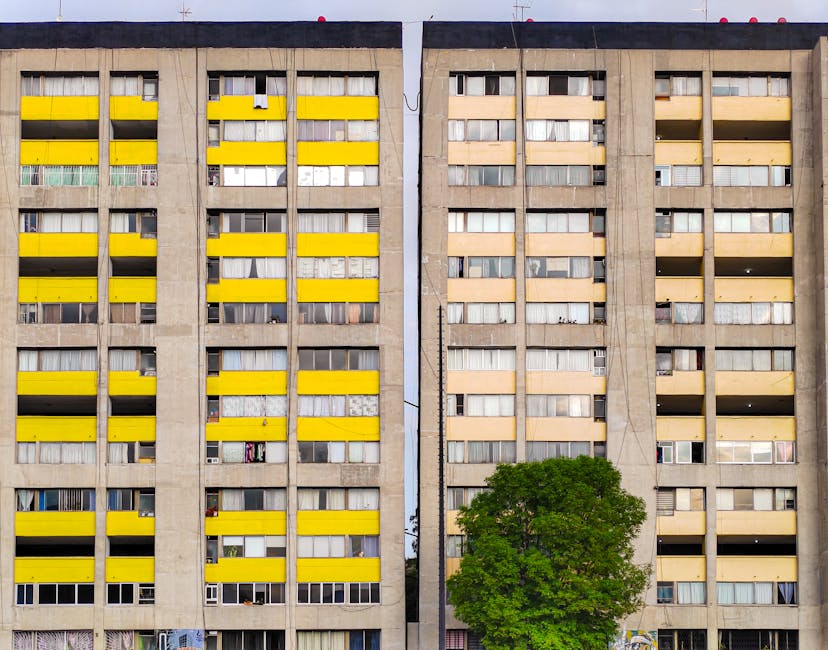 A high-rise residential building with a concrete facade displaying multiple floors with windows and balconies, some of which are closed or open. Bright yellow window blinds are visible on several floors, contrasting with the neutral grey exterior. The balconies vary in appearance, with a few showing laundry or furniture, and some windows have curtains or blinds partially drawn. In front of the building, a lush green tree stands, adding natural contrast to the urban structure. The overall scene is well-lit with natural daylight, highlighting the building's details. This image relates to residential properties and may be used by Kennington Carpet Cleaning to illustrate the importance of surface cleaning and maintenance for multi-story flats in Vauxhall and Kennington, supporting their end-of-tenancy cleaning services.
