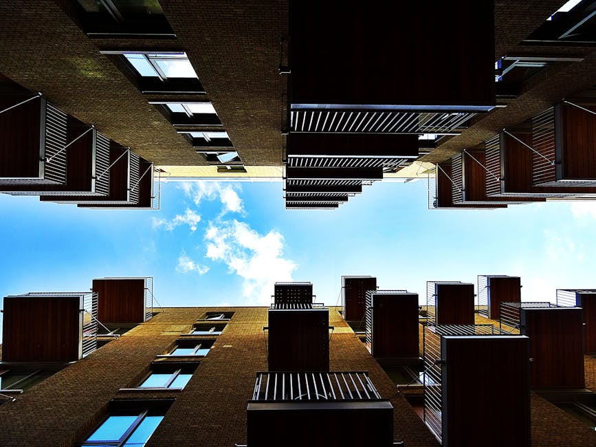 Looking up from the courtyard of a multi-story residential building, the view captures the facades with balconies fitted with metal railings and wooden paneling. The exterior surfaces are clean and well-maintained, with large windows reflecting the bright blue sky and scattered white clouds above. The building's brickwork appears tidy, and the communal outdoor space is free of debris. Natural daylight illuminates the scene, highlighting the neatness and quality of the building's exterior. This setting suggests a focus on building maintenance and surface cleaning, exemplifying professional care for residential property exteriors, aligned with services provided by Kennington Carpet Cleaning as part of their cleaning expertise.