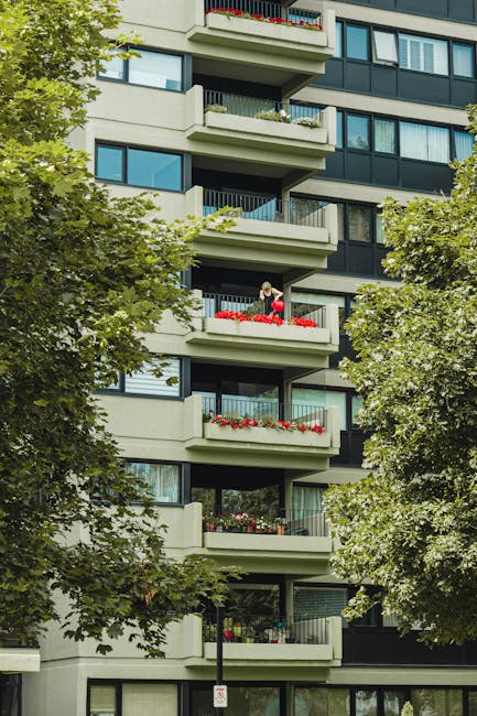 A high-rise residential building with a concrete facade displaying multiple floors with windows and balconies, some of which are closed or open. Bright yellow window blinds are visible on several floors, contrasting with the neutral grey exterior. The balconies vary in appearance, with a few showing laundry or furniture, and some windows have curtains or blinds partially drawn. In front of the building, a lush green tree stands, adding natural contrast to the urban structure. The overall scene is well-lit with natural daylight, highlighting the building's details. This image relates to residential properties and may be used by Kennington Carpet Cleaning to illustrate the importance of surface cleaning and maintenance for multi-story flats in Vauxhall and Kennington, supporting their end-of-tenancy cleaning services.