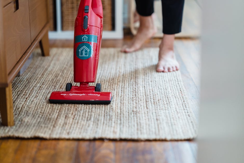 A person vacuuming a beige textured rug in a domestic setting, with their bare feet visible on the edge of the rug. The vacuum cleaner, which is red with a light blue control panel and a brush head labeled 'easy lightweight cleaning,' is positioned on the carpeted area, supported by a wooden floor. In the background, there is a wooden piece of furniture, possibly a sideboard or cabinet, under warm ambient lighting. The scene demonstrates surface cleaning and deep cleaning processes aimed at maintaining hygiene and cleanliness in a home environment, as offered by Kennington Carpet Cleaning.