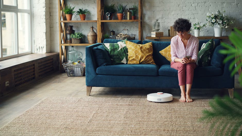 A woman sitting barefoot on a blue sofa in a living room with a beige rug, using a smartphone. The room features a wooden shelving unit with potted plants, decorative items, and books positioned against a textured gray wall. A white robotic vacuum cleaner is on the rug near her feet. Natural light filters in through large windows on the left side, illuminating the space. The scene highlights domestic cleaning and surface maintenance with a focus on modern, tidy interior design, as emphasized by Kennington Carpet Cleaning in their guide for local homes.