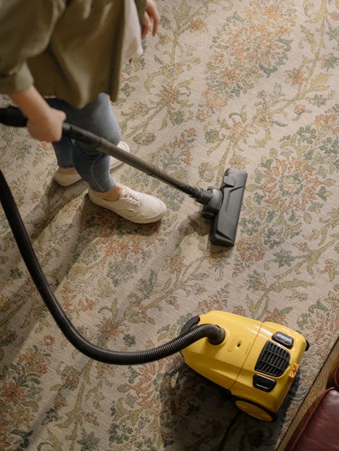 A person vacuuming a beige textured rug in a domestic setting, with their bare feet visible on the edge of the rug. The vacuum cleaner, which is red with a light blue control panel and a brush head labeled 'easy lightweight cleaning,' is positioned on the carpeted area, supported by a wooden floor. In the background, there is a wooden piece of furniture, possibly a sideboard or cabinet, under warm ambient lighting. The scene demonstrates surface cleaning and deep cleaning processes aimed at maintaining hygiene and cleanliness in a home environment, as offered by Kennington Carpet Cleaning.