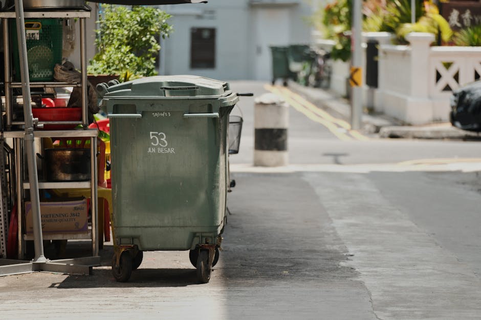 A green wheeled waste disposal bin situated on the sidewalk outside a building in a residential area. The bin is positioned next to a metal rack and partially in front of a storefront, with gardening tools and miscellaneous items visible nearby. The surface of the sidewalk appears clean and dry, with a section of asphalt paving. In the background, a street with bollards, parked cars, and greenery from shrubs and trees can be seen, illuminated by natural daylight. This setting illustrates waste management related to local council regulations as discussed on the Lambeth Council rules for waste disposal after cleaning page, with Kennington Carpet Cleaning’s involvement in maintaining hygiene and cleanliness standards.
