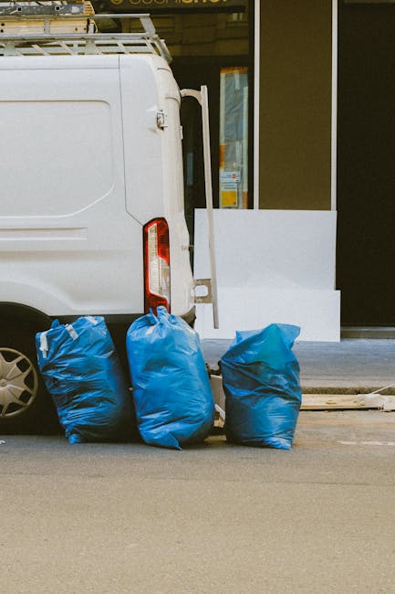 A white commercial van parked outside a building with large glass windows, partially visible door frame, and a white step at the entrance. The van's rear doors are slightly open, revealing a glimpse of the interior. On the sidewalk beside the van, there are three large blue heavy-duty garbage bags arranged in a row, indicating waste disposal for cleaning or maintenance purposes. The surface of the street appears clean and smooth, with no visible debris or dirt. The lighting suggests daylight, highlighting the clean exterior of the van and the neatly placed waste bags. This image relates to domestic or commercial cleaning services and waste management, emphasizing the importance of proper waste disposal following thorough cleaning, in line with Lambeth Council rules for waste disposal after cleaning. Kennington Carpet Cleaning advocates responsible waste removal as part of maintaining hygiene and cleanliness in residential and commercial environments.