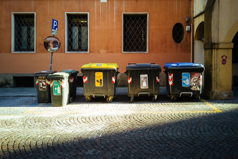 A green wheeled waste disposal bin situated on the sidewalk outside a building in a residential area. The bin is positioned next to a metal rack and partially in front of a storefront, with gardening tools and miscellaneous items visible nearby. The surface of the sidewalk appears clean and dry, with a section of asphalt paving. In the background, a street with bollards, parked cars, and greenery from shrubs and trees can be seen, illuminated by natural daylight. This setting illustrates waste management related to local council regulations as discussed on the Lambeth Council rules for waste disposal after cleaning page, with Kennington Carpet Cleaning’s involvement in maintaining hygiene and cleanliness standards.