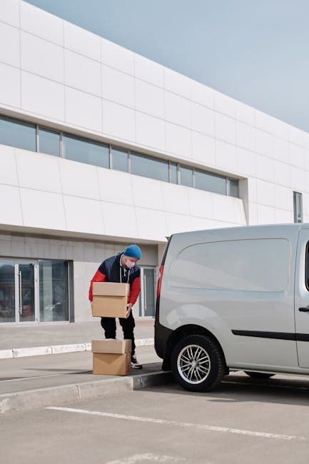 A white commercial van parked on a street outside a building with large glass windows, partially visible door frames, and a white step. The van's rear door is closed, and three blue recycling bags are placed on the ground near the back wheel, containing waste. The street surface is asphalt, and the scene is well-lit with natural light, highlighting the clean, smooth surfaces of the van and the neat placement of the bags. This setting reflects a typical urban environment where Kennington Carpet Cleaning carries out domestic or commercial cleaning services, emphasizing hygiene and proper waste disposal as part of the cleaning process. The background includes minimal visible details but suggests a tidy, organized exterior consistent with professional cleaning standards.