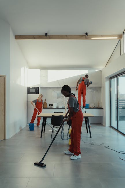 A team of three professional cleaners wearing red aprons and casual clothing performing surface cleaning tasks in a modern, minimalist kitchen with light-colored tiled floors, white walls, and large glass sliding doors. One cleaner is using a vacuum cleaner on the floor, while another is mopping the tiled surface near the countertop. The third cleaner is working on the upper wall near a window. The kitchen features stainless steel appliances, a white countertop with integrated sink, and subtle wooden ceiling accents, all illuminated by natural light from the large glass doors. The space appears clean, well-maintained, and organized, reflecting thorough deep cleaning and surface sanitisation practices by Kennington Carpet Cleaning.