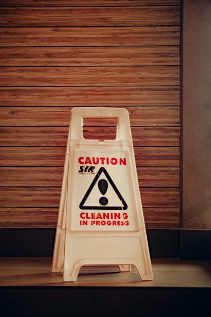 A white plastic 'Caution Cleaning in Progress' sign with a black triangular warning symbol and red text is placed on a tiled floor near a wooden-paneled wall in an indoor environment. The sign indicates ongoing cleaning activities. The lighting is moderate, highlighting the sign and the textured wood surface of the wall, emphasizing the caution warning related to sanitation and surface cleaning procedures performed by Kennington Carpet Cleaning, a professional cleaning company. The setting suggests a commercial or public space undergoing cleaning or maintenance to ensure hygiene and cleanliness.