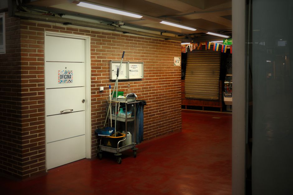 A team of three professional cleaners wearing red aprons and casual clothing performing surface cleaning tasks in a modern, minimalist kitchen with light-colored tiled floors, white walls, and large glass sliding doors. One cleaner is using a vacuum cleaner on the floor, while another is mopping the tiled surface near the countertop. The third cleaner is working on the upper wall near a window. The kitchen features stainless steel appliances, a white countertop with integrated sink, and subtle wooden ceiling accents, all illuminated by natural light from the large glass doors. The space appears clean, well-maintained, and organized, reflecting thorough deep cleaning and surface sanitisation practices by Kennington Carpet Cleaning.