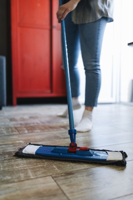 A person wearing blue jeans and white socks is using a flat mop with a blue handle and microfiber cleaning pad to perform surface cleaning on a wooden floor in a domestic kitchen setting. The floor appears clean and well-maintained, with visible wood grain. In the background, there is a red cupboard, part of a white refrigerator, and nearby natural light illuminating the area, highlighting the hygiene and maintenance efforts associated with professional cleaning by Kennington Carpet Cleaning, which explains the importance of paying the real cost of affordable cleaning options in Kennington.