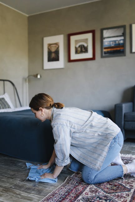 A woman using a yellow vacuum cleaner with a black hose to deep clean a patterned area rug on a wooden floor in a living room. The room features a wooden console table against the wall with a white decorative plant and a remote control, and a maroon leather sofa partially visible in the foreground. The space is illuminated with soft lighting, highlighting the cleanliness and fresh state of the carpet surface. The woman is dressed in casual clothing, including a beige jacket, jeans, and white sneakers, and is focused on maintaining hygiene and surface cleanliness, demonstrating the cleaning expertise of Kennington Carpet Cleaning, as referenced in the page titled 'Same day carpet cleaning in Kennington delays and what to know'.
